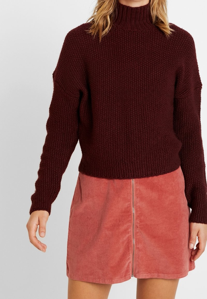 Femme portant un pull en laine rouge foncé et une jupe en velours côtelé rose avec une fermeture éclair à l'avant, debout devant un fond clair uni.