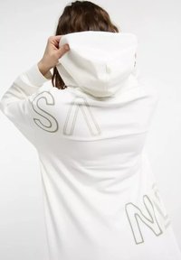 Person adjusts the hood of a white sweatshirt displaying large, stylized letters on the back. Background is plain and light-colored.
