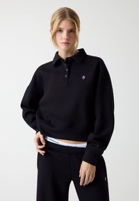 Zwarte cropped sweatshirt met kraag, drieknoops steek en geborduurd logo. Gecombineerd met zwarte broek met witte tailleband waarop branding staat.