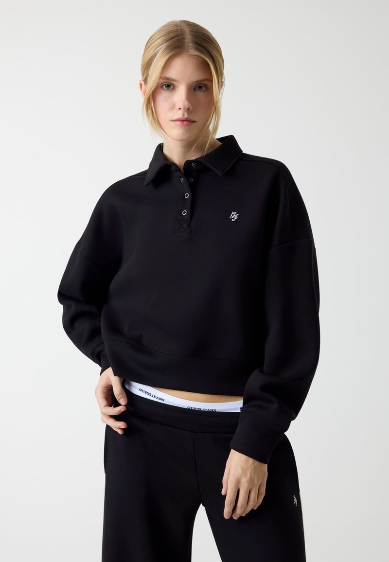 Zwarte cropped sweatshirt met kraag, drieknoops steek en geborduurd logo. Gecombineerd met zwarte broek met witte tailleband waarop branding staat.