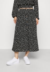 Mustat midihame valkoisilla abstrakteilla täplillä, jossa on nappikiinnitys edessä, joustava vyötärö ja valkoiset tennarit jalassa.