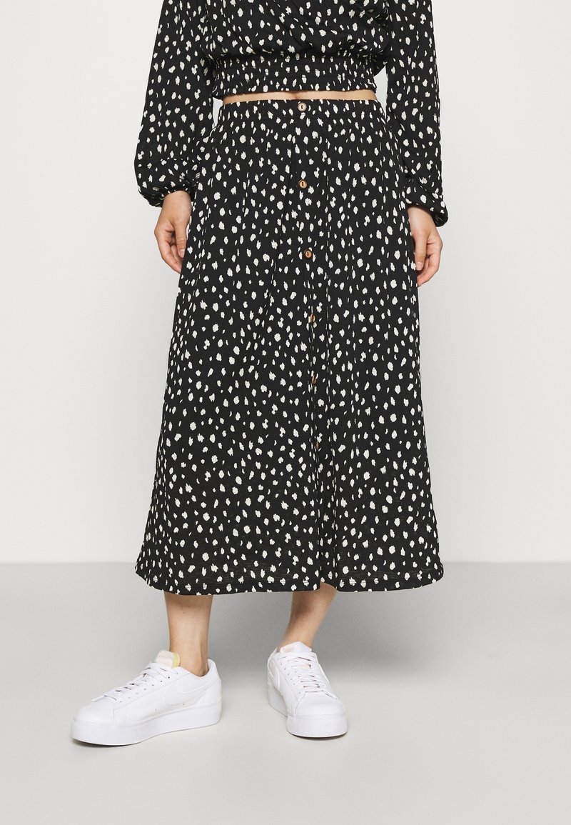 Mustat midihame valkoisilla abstrakteilla täplillä, jossa on nappikiinnitys edessä, joustava vyötärö ja valkoiset tennarit jalassa.
