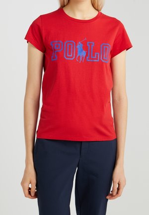 Mujer vistiendo una camiseta roja con texto azul "POLO" y un logo de jugador de polo, combinado con pantalones azul oscuro, sobre un fondo liso.