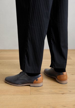 Des chaussures noires et grises avec une texture lisse, présentant un accent en cuir marron au talon. Portées avec un pantalon noir à rayures.
