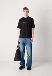 Svart bomull T-skjorte med "Calvin Klein" logo, kombinert med lyseblå revet bukser, svarte sko og en svart totebag, stående på en enkel bakgrunn.