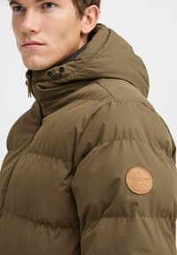 Brown puffy jacket with a hood, featuring a leather logo patch on the sleeve and a textured fabric. Zipper closure visible at the front.