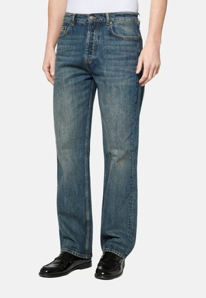 Man draagt vervaagde rechtbenige blauwe jeans, zwarte leren loafers en een wit shirt tegen een witte achtergrond.