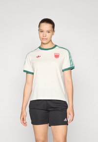 Cream short-sleeve shirt with green collar and sleeves, featuring a red shield logo and small red adidas emblem, paired with black shorts.