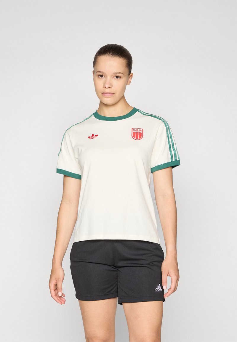Cream short-sleeve shirt with green collar and sleeves, featuring a red shield logo and small red adidas emblem, paired with black shorts.
