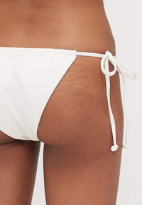Bela spodnjica bikinija z teksturiranim vzorcem, nastavljive vrvice na straneh, z gladkim zaključkom in minimalnim oblikovanjem pokritosti.