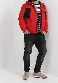 Rød jakke med isolering og sorte detaljer, lodrette lynlåslommer, stribet skjorte under, sorte jeans og beige sneakers.