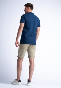 Laivaston värinen polo-paita, beige käännettävät shortsit, valkoiset tennarit laivaston yksityiskohdilla. Sileä kangas, rento istuvuus, minimaalinen yksityiskohtaisuus. Takana näkyvä mies.