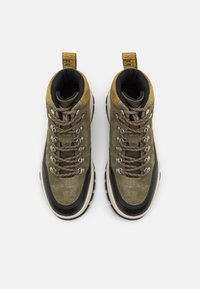 Wysokie buty trekkingowe w oliwkowym zamszu i czarnym gumie. Posiadają brązowe sznurówki, metalowe oczka oraz żółtą pętelkę z tyłu do łatwego zakupu.