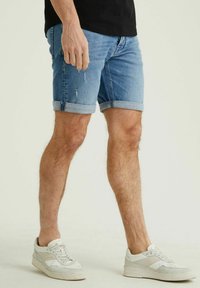 Džinsiniai šortai, šviesiai mėlynos spalvos su apvartu ir nusidėvėjimo detalėmis. Sujungti su baltais sportiniais bateliais, turinčiais tekstūrinių akcentų.