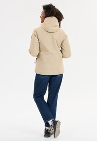 Beige hoodie-jack met een soepele textuur, een getailleerde silhouette en een verstelbare trekkoordzoom, gecombineerd met blauwe spijkerbroek en robuuste sneakers.