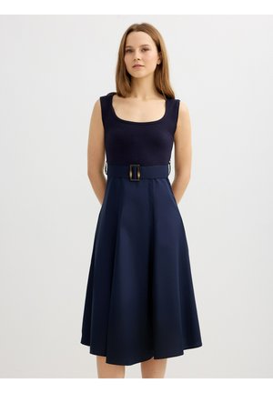 Mujer con vestido azul marino sin mangas con escote cuadrado y cinturón ancho con hebilla rectangular, de pie sobre fondo liso.