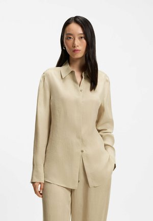 Pitkähiuksinen nainen, jolla on beige napillinen paita ja vastaavat housut, seisoo valkoista taustaa vasten pitäen yhtä kättään taskussa.