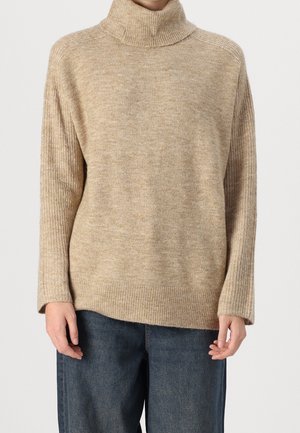 Pull à col roulé beige en maille avec des poignets et un bas côtelés, porté avec un jean bleu foncé.