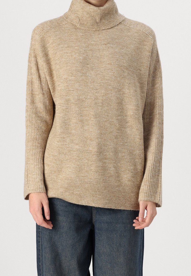 Pull à col roulé beige en maille avec des poignets et un bas côtelés, porté avec un jean bleu foncé.