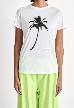 Personne portant un T-shirt blanc avec un imprimé de palmier noir et un pantalon vert fluo, debout devant un fond blanc uni.