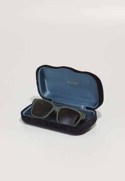 Ochelari de soare pătrați verzi într-un toc din catifea neagră și albastră, având branding-ul auriu "GUCCI" pe interiorul capacului.