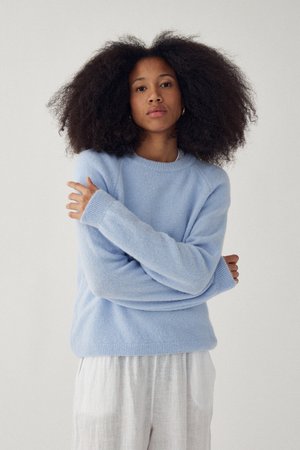 Žena s objemnými kučeravými vlasmi, oblečená v svetlomodrom svetri a bielych nohaviciach, stojaca so založenými rukami pred jednoduchým pozadím.