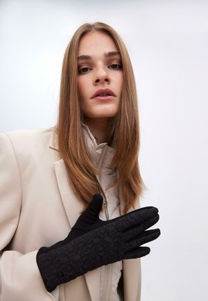 Guantes negros con un patrón texturizado, que presentan un puño largo y un acabado de tela suave, utilizados por una persona con un abrigo de color claro.