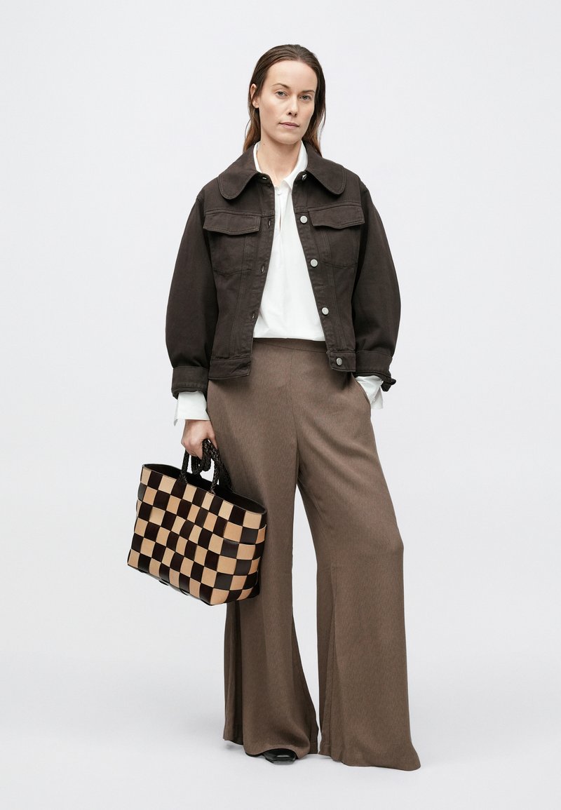 Veste en jean brun foncé, chemise blanche et pantalon large brun. Tenant un sac fourre-tout noir et beige à carreaux avec des poignées courtes.