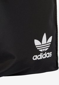 Černá tkaninová taška s bílým logem Adidas a ikonickým designem trefoil. Hladká textura s prošitím na okrajích a bez viditelných kovových částí.