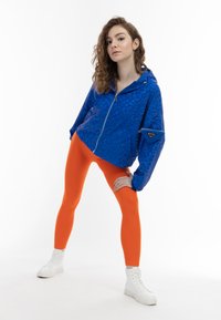 Giacca blu a fantasia con cappuccio e chiusura con zip, abbinata a leggings arancioni aderenti e sneakers bianche alte.