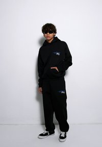 Czarny zestaw z kapturkiem i spodniami dresowymi z kieszenią z przodu. Niebieski nadruk graficzny na obu elementach. Uzupełniony czarnymi trampkami z białymi akcentami.