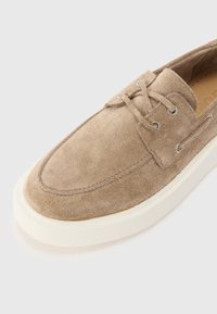 Beige velúr cipő kerek orral, fűzős kialakítással, ezüst fűzőlyukakkal és fehér gumitalppal. A texturált felszín részletességet ad hozzá.