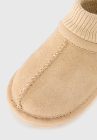 Chaussure en suède beige avec une cheville en maille côtelée. Elle présente un bout rond et des coutures visibles le long de la couture et de la semelle. Texture douce, design décontracté.