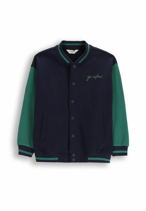 Granatowa kurtka z zielonymi rękawami, ryflowanym kołnierzem i mankietami. Posiada zatrzaski i kieszenie; haftowane hasło "idziesz na odkrywanie!" z przodu.