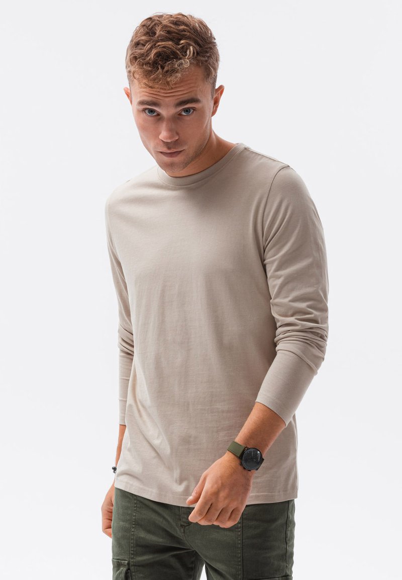 Beige Langarmshirt mit Rundhalsausschnitt und enganliegenden Ärmeln, kombiniert mit grünen Cargo-Hosen und einer schwarzen Uhr. Glatte Textur.