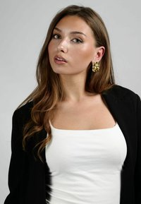 Pendientes de oro texturizados; blusa blanca ajustada; chaqueta negra. Cabello largo, ondulado y castaño; maquillaje neutro con toques naturales.