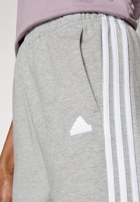 Šedé bavlněné tepláky se třemi bílými pruhy po stranách a malým bílým logem Adidas na oblasti přední kapsy.