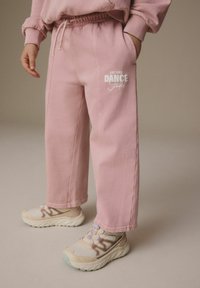 Rosa sweatpants med resårband i midjan har texten "DANCE Studio" i vitt. Bärs tillsammans med beige och rosa sneakers med strukturerad detaljering.