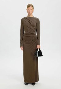 Femme en robe maxi marron à manches longues froncées tenant un petit sac à main noir, portant des chaussures noires, debout devant un fond blanc.