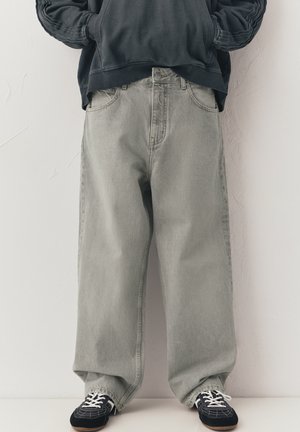 Osoba v volných šedých džínách, černých teniskách s bílými tkaničkami a černé mikině, stojící před jednobarevnou bílou zdí.