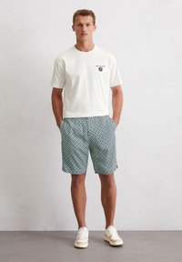 White cotton t-shirt with black logo, paired with green patterned shorts. Model wears white sneakers with a textured sole.