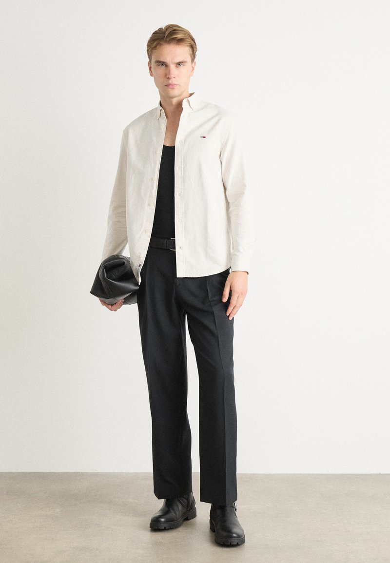 Camisa de botón beige claro, camiseta de tirantes negra, pantalones negros y botas negras. Sosteniendo un bolso de mano negro. Diseño minimalista con líneas limpias.