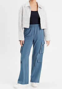 Geknipte, gestructureerde witte blazer met blauwe accenten; zwarte aansluitende tanktop; losse blauwe cargobroek met zijzakken; witte sneakers.