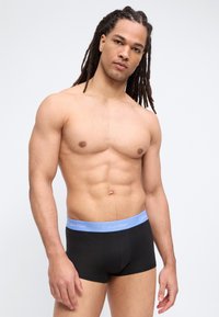 Άνδρας χωρίς μπλούζα με μακριά dreadlocks, φοράει μαύρο εσώρουχο Calvin Klein με ανοιχτό μπλε ζωνάκι στη μέση, μπροστά σε απλό λευκό φόντο.