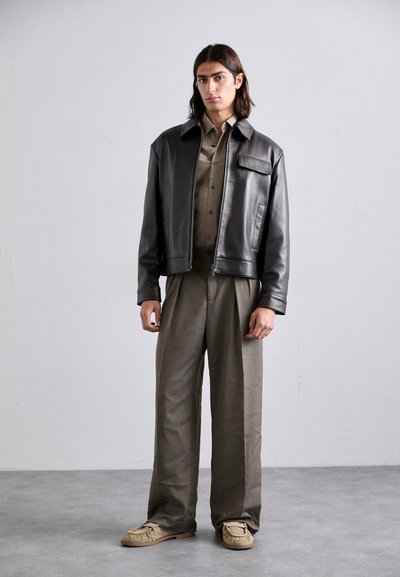 Black leather jacket with a collar and front pocket, paired with an olive green shirt and light gray trousers. Beige slip-on shoes.