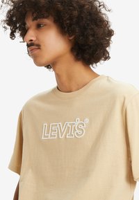 Beezist puuvillane T-särk, millel on valge õmmeldud logo "LEVI'S®". Lühikesed varrukad ja ümar kaelus, sujuva tekstuuriga.