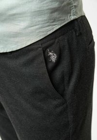 Svarta byxor med en slät textur, med en sidoficka som har en diskret broderad logotyp. Kombineras med en ljusgrön skjorta ovanför.