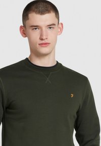 Sweat-shirt vert foncé à encolure ras du cou, avec un détail de point croix subtil sur l'épaule et un petit logo jaune sur la poitrine.