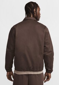Brun letvægtsjakke med ståkrave, elastiske ærmer og en kantet bund. Glat tekstur, simpelt design og ingen synlige mønstre.