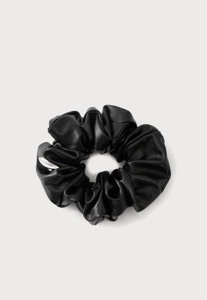 Chouchou noir brillant en tissu similicuir froncé avec une petite étiquette argentée sur fond gris clair.